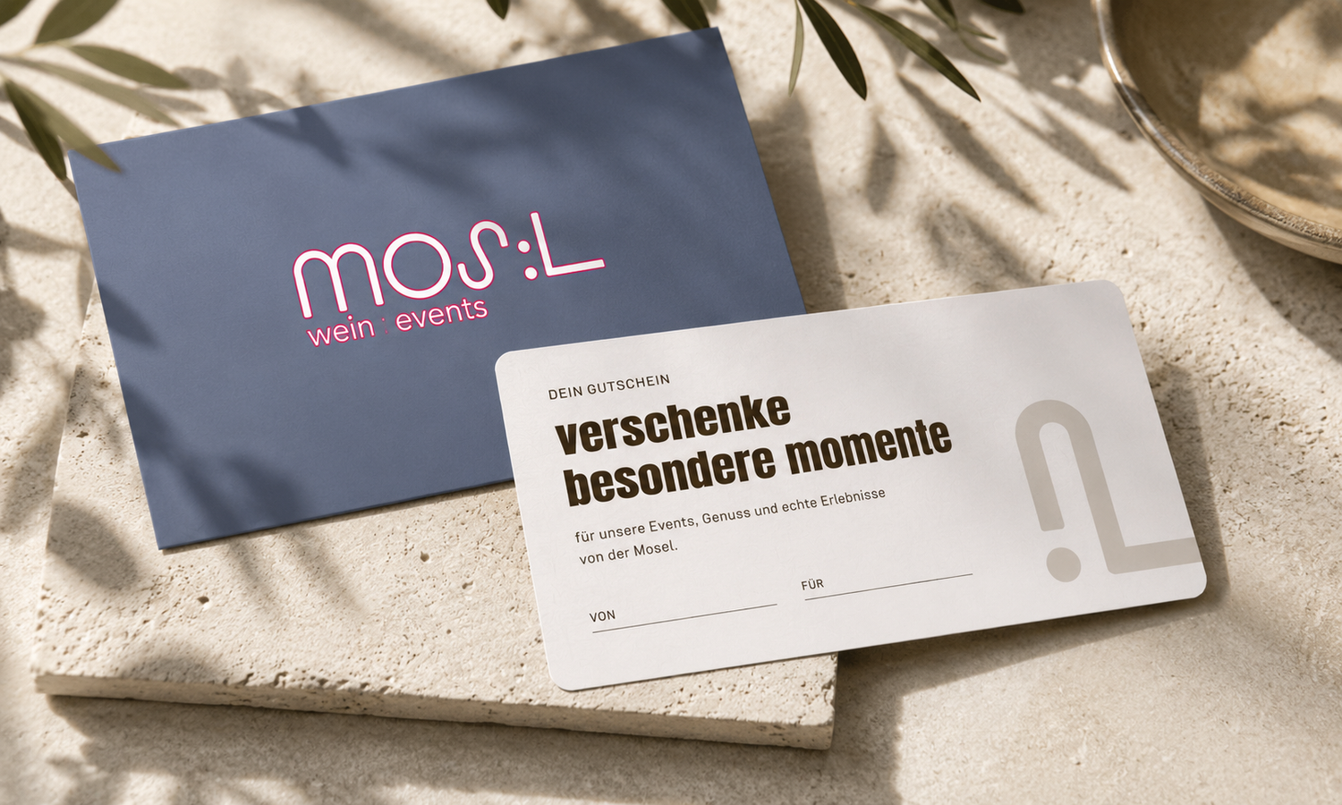 Stilvoll inszenierter MOS:L Gutschein mit Rückseite und passendem Umschlag auf heller Steinfläche, umgeben von warmem Sonnenlicht, Olivenzweigen und mediterranen Details – als Geschenk für Wein, Events und Genussmomente.