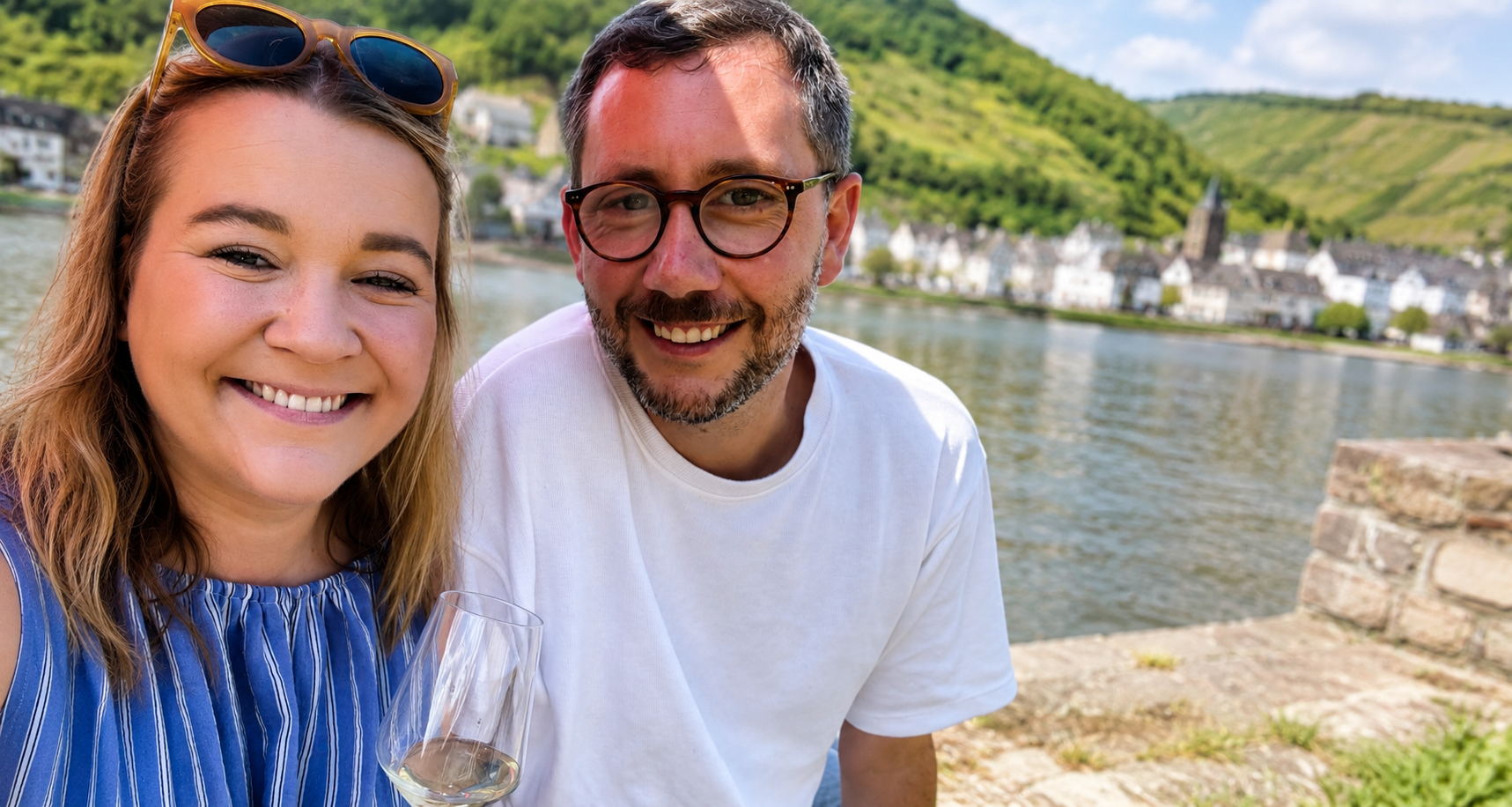 Sympathisches Porträt von zwei Personen am Ufer der Mosel mit einem Glas Weißwein in der Hand, vor der Kulisse von Fluss, Weinbergen und einem typischen Moselort.